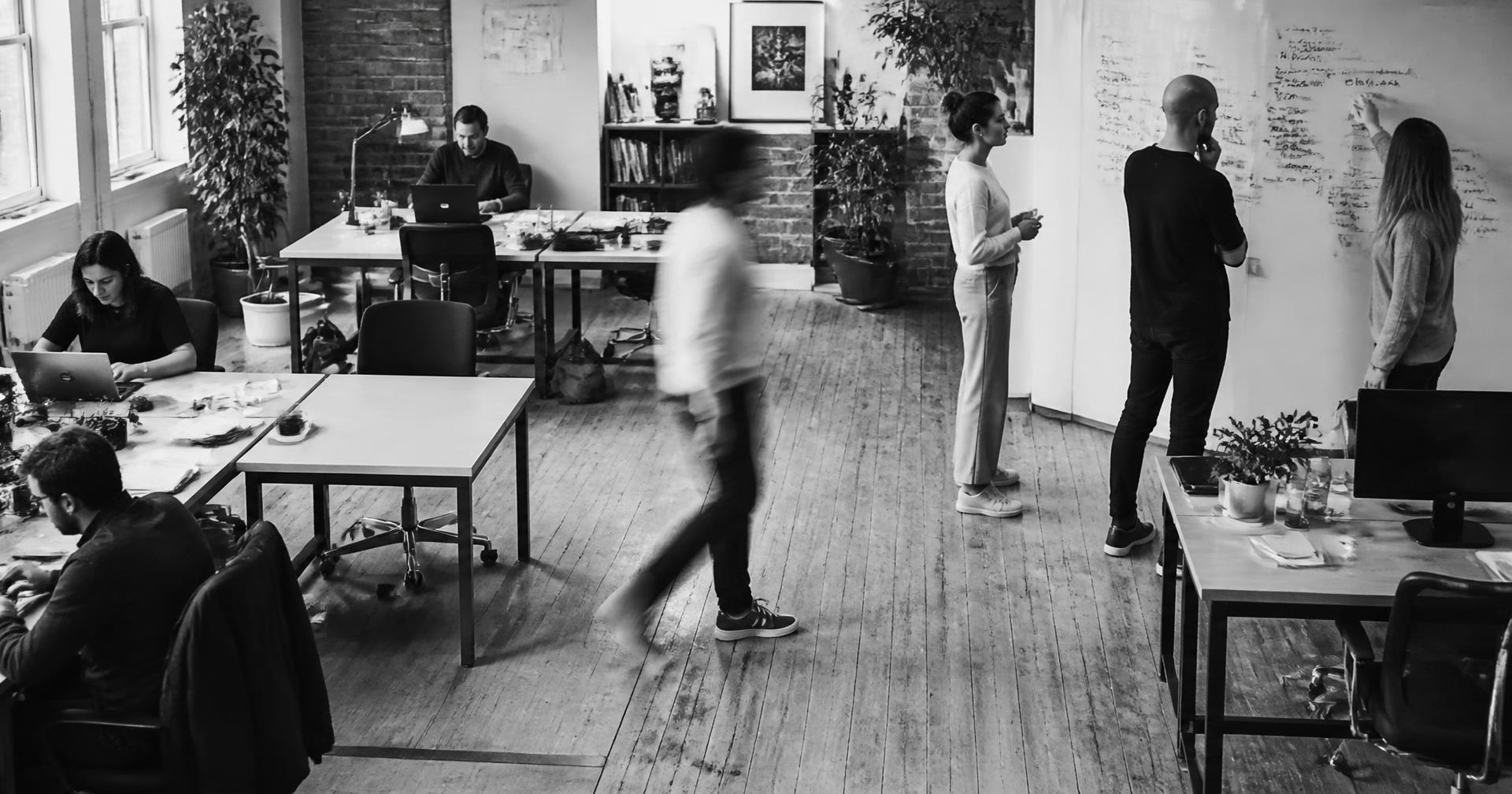 A busy agency workspace: people at desks, a discussion at a whiteboard, someone walking through